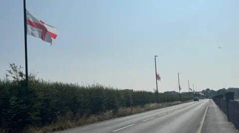 BBC An A road which is lined with a hedge to the left, along every lamppost on the street is a St George's Cross flag