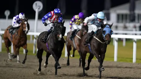 Two jockeys on horses, and two behind them, are racing on a sandy track at Chelmsford City Racecourse at night-time. The white fencing is visible and a building in the background out of focus.