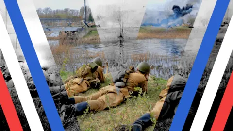 A photo showing men wearing World War Two military uniforms lying on their stomachs in the grass in front of a river. On the other side of the river plumes of smoke are erupting into the cloudy sky