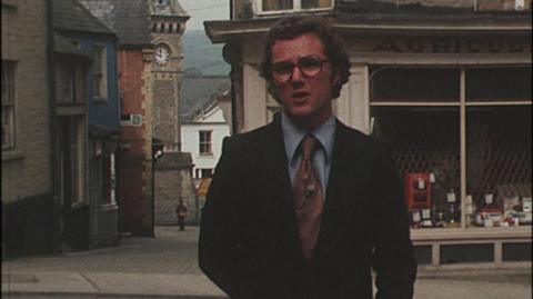 The presenter, wearing a suit and tie, standing in a street on Hay-on-Wye in front of a shop.
