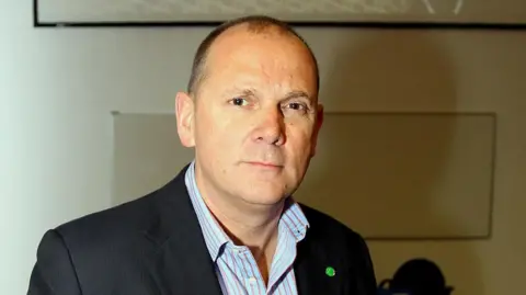 PA Jim Gamble, wearing a black blazer and a striped shirt