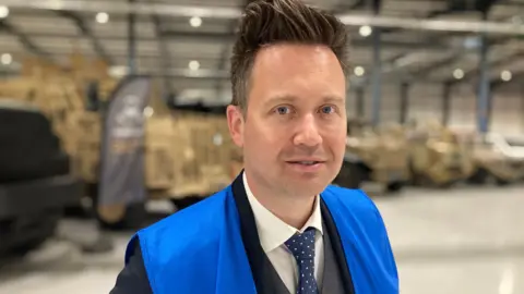 A man wearing a suit and bright blue vest looks into the camera. Various machinery can be seen blurred in the background.