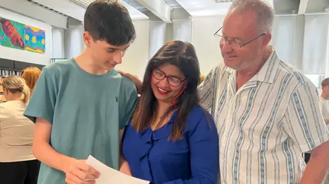 Ethan is wearing a teal T-shirt and he is holding a white piece of paper. His mother and father are looking on pleased.