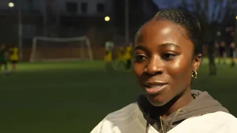 TV screenshot of Sakinah Boateng, a former footballer. She is standing by the edge of a football pitch on which a training session is taking place. It is dark and the pitch's lights are on. She is wearing a pale brown hoodie and a cream top