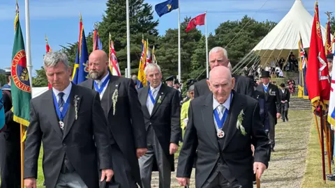 5 men all in dark suits (with a stretch of green herb pinned to their chest) walk down a procession of flags 