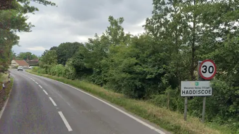 Google A road with broken central white lines. A sign to the right says "Waveney Valley Village, Haddiscoe", with a "30" sign above it. The road is edged with grass and trees.