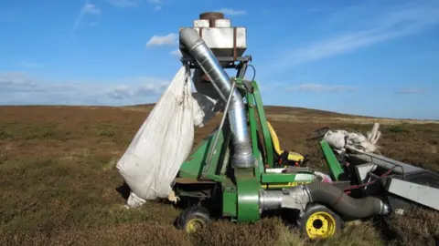 A piece of green machinery with a white sack at one end, held up by a green metal arm on wheels