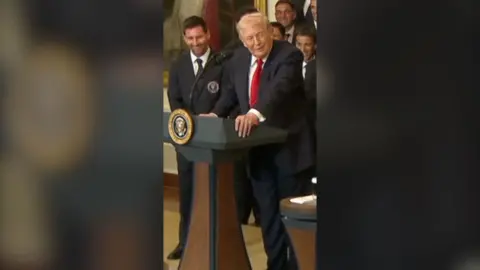 Lionel Messi (left) stands next Donald Trump (right) who is behind a podium. Both are suited