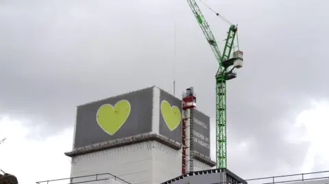 Grenfell Tower partially covered in grey cladding with green heart symbols, as a crane stands beside the building during deconstruction work.