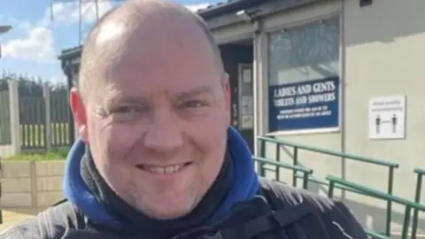 A picture of a man who smiles at the camera. He is wearing black clothing as well as an item with a blue collar.