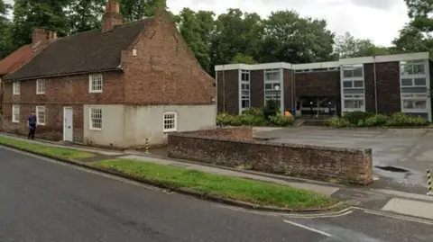 Two contrasting buildings face an empty asphalt road: In the foreground on the left behind a strip of grass is an 18th Century steep roofed building with small windows. To the right of it is a car park, at the rear of which is a 1960s brick office building.
