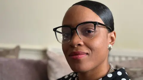 Dainya in a black and white polka dot blouse. She wears a necklace, earrings and glasses. She is sat on a sofa looking at the camera.