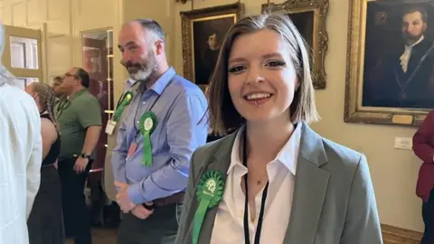 A woman with neck-length brown hair and a lip piercing smiles at the camera. She is wearing a white blouse under a grey jacket with a green rosette pinned to her lapel.