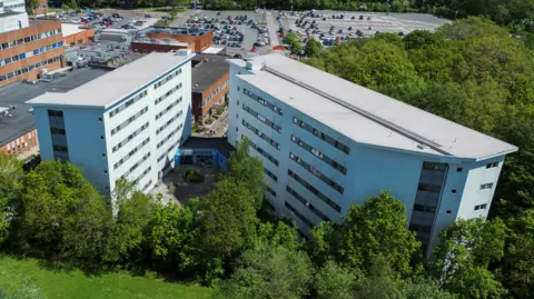 A drone view of Arrowe Park Hospital. Two multi-storey blue/grey buildings loom above trees