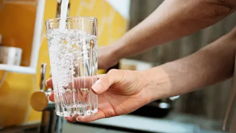 Getty Images A woman fills a clear drinking glass with water under the tap.