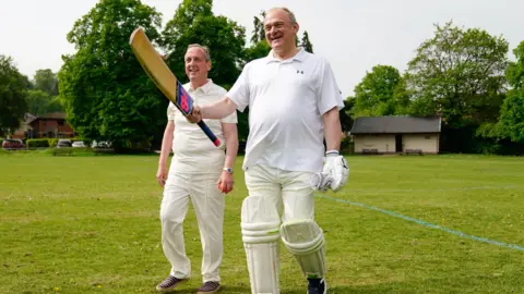 PA Media Liberal Democrat Leader Sir Ed Davey (right) coming out to bat with Lib Dem group leader councillor Ian Thorn. They are wearing cricket whites and Sir Ed has a cricket bat in his hand which he is holding in the air.