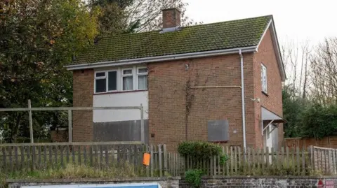 A two-storey house made out of brick that has been partially boarded up. It has a fence in front of it and a tree to the side. The surrounding scene is overgrown and the house has clearly not been used for some time.