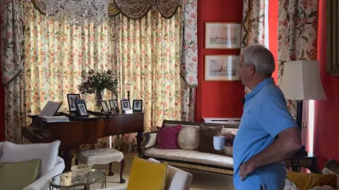 Major Beaumont stands in a grand room with a coffee cup in his hand, wearing a blue polo top. He is looking at a brown piano, with portaits of royal visitors standing on it.