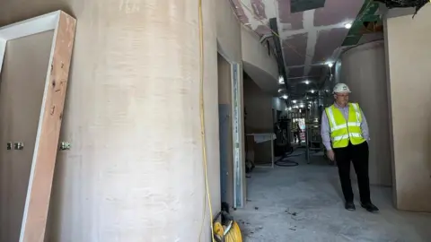 NHS A corridor in a diagnostic centre where construction work is under way. Plaster can be seen on the walls and a man in a hard hat and high vis jacket is on the right of the photo 