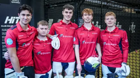 Queen's College Five young men in dark pink cricket shirts standing in front of a net, holding their gloves and helmets and smiling to camera