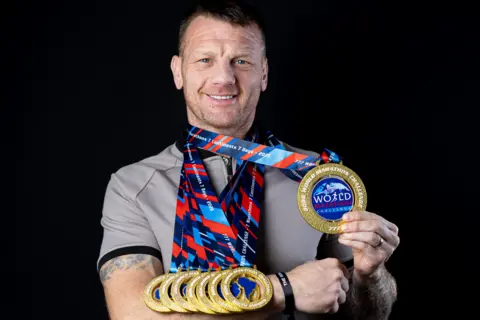World Marathon Challenge A grinning Bobby, who is wearing a grey t-shirt, balances six of his medals on the right forearm and holds aloft another with his left hand