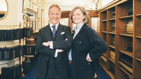 Redmayne 1860 Tailoring Claire and Tom Mahon standing side-by-side and smiling in a room filled with shelves stocking different fabrics. Fabric samples are hung from rails down one of the walls. They are both dressed in formalwear attire.