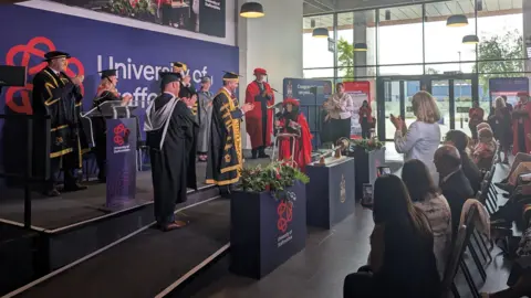 People in formal wear clap as a woman in a red gown and cap uses a walking aid to cross a stage.