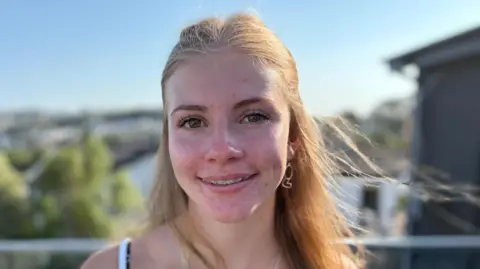 Lola is smiling into camera, her fixed brace is visible on her upper teeth. She has large dark eyes and long fair hair tied back from her face. The background is blurred but you can see houses and greenery in the distance.