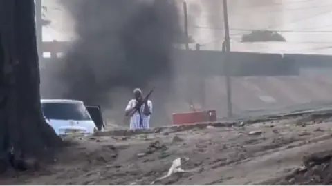TikTok A man in civilian clothes holds a rifle, standing beside a saloon car. Black smoke rises behind him.