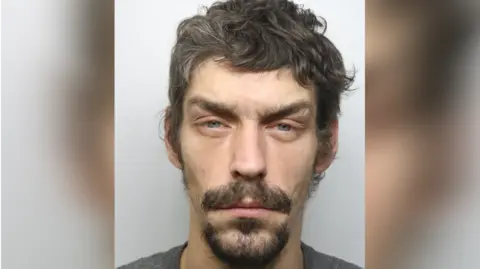 A custody photo of a man with dark hair, a dark beard, and moustache
