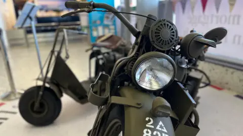 A close up of the front of a Harley-Davidson WLA, it is camouflage green. In the background a a Cushman Airborne Scooter. The background is blury 