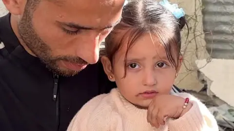 Julia Abu Warda, aged three, sits on her father's knee, as he looks down. There is a wary expression in her brown eyes. She is dressed in a peach-coloured jumper, with her hair in two buns tied with blue bobbles.  