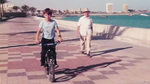 The Buckley Society To the left a child is on a bike, and a man to the right wearing all white. They are walking along a brick coastal path, the sea can be seen the right of them. 