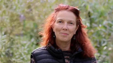 Kai Hilton Henri Brocklebank, a woman with long red hair, wears a jersey and a body warmer and stands in a field.