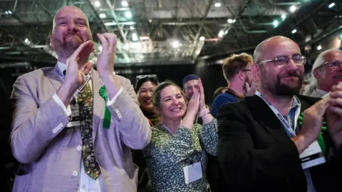 PA Men and women, some wearing suits, but all with green rosettes clap and cheer inside an arena