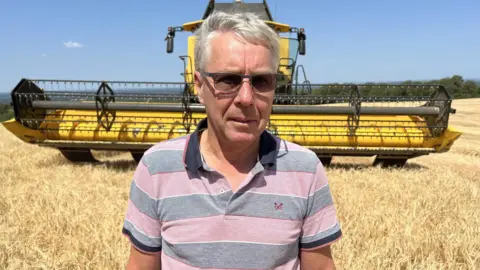 Leanne Rinne/BBC A farmer standing in a field of barley in front of a large yellow combine harvester
