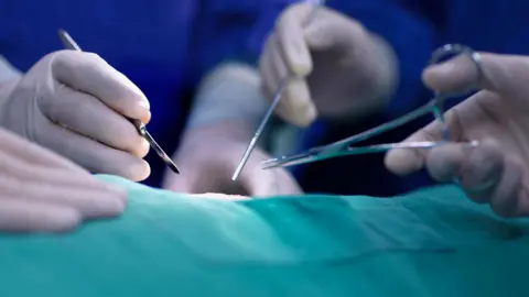 Getty Images is a public library image, which appears closely from several hands in surgical gloves that carry medical tools including scissors and scalpel, near part of the patient protected by vision by green tissue.