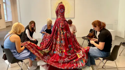 Kirstie Macleod A red dress on a mannequin with women sat on chairs around the dress and embroidering it.