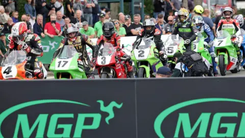 MANX GRAND PRIX Riders line up on their motorbikes on he start line. There is a sea of spectators behind them and boards with the MGP logo on them in front of them.