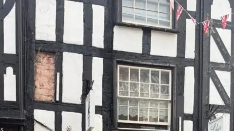 Glenn Jenkinson-Deakin A close-up of rendered timber frame on a 17th Century building, painted black and white. A section of timber has fallen away, leaving exposed brickwork. A sash window can be seen partially open, with lace curtains, and Union Jack bunting.