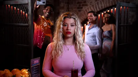 BBC/Jack Barnes/Kieron McCarron A young woman with bleached blonde shoulder length hair wears a pink top and holds a drink in a glass with straw held in two hands at waist level. Behind her a group of friends takes a selfie in a dimly lit bar or nightclub.ford, who plays Anna Knight