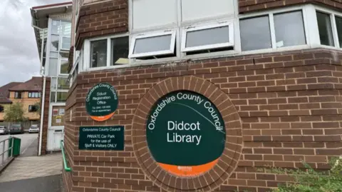The front entrance of Didcot Library. It is a red brick building with a disabled ramp on the left and a circular green sign directing people inside.