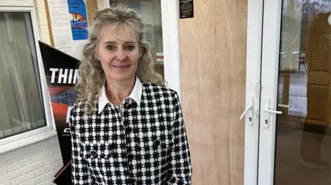 Director of golf at the West Malling club Kate Brett stands in front of a boarded-up door. She is wearing a black and white top with a white collar and has long, wavy blonde hair. There are club signs and posters behind her.
