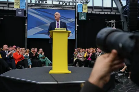 PA Media John Swinney standing at a bright yellow podium on a small stage inside an indoor venue, speaking to an audience seated around the stage, with a large blue-and-white backdrop behind the podium and a camera lens visible in the foreground.”