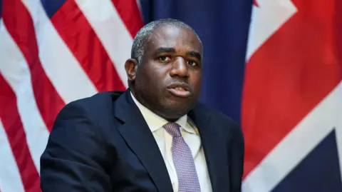 Reuters David Lammy looks ahead while sitting down in a dark blue suit and a patterned tie made up of small white and pink checks. The flags of the US and UK can be seen behind him.