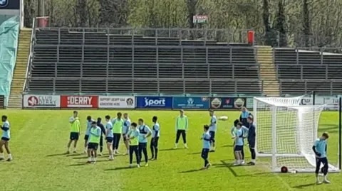 Shows 19 male footballers in a training session at a stadium