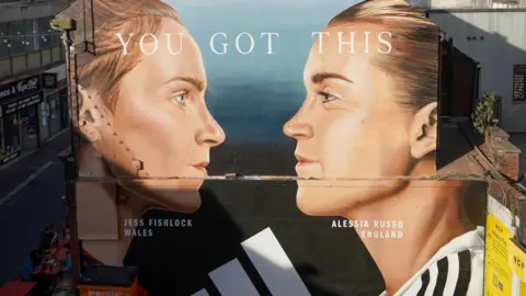 A mural painting on a side of the wall featuring the two football players, Jessica Fishlock and Alessia Russo. It shows their faces in profile, facing each other. Their hair is tied back and their football shirts are just visible at the bottom of the mural. 