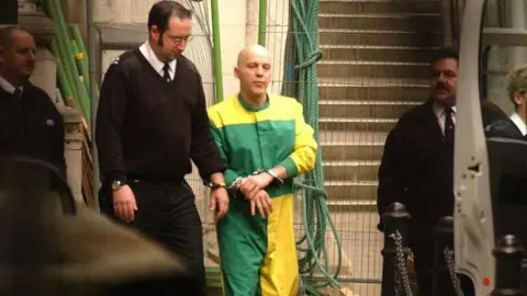 PA Media A man with a closely shaven head and a green and yellow prison suit is being led in handcuffs by a prison officer. Other security staff can be seen to either side of him.