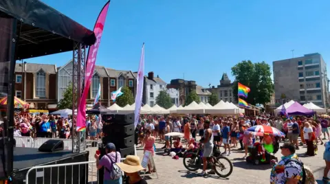 West Northamptonshire Council A large group of people in a market square at a Pride event with flags and stalls. A stage is to the left. Buildings surround the square.