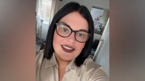 Naomi Bedding A woman with shoulder-length dark brown hair and brown eyes wearing tortoiseshell glasses taking a selfie.